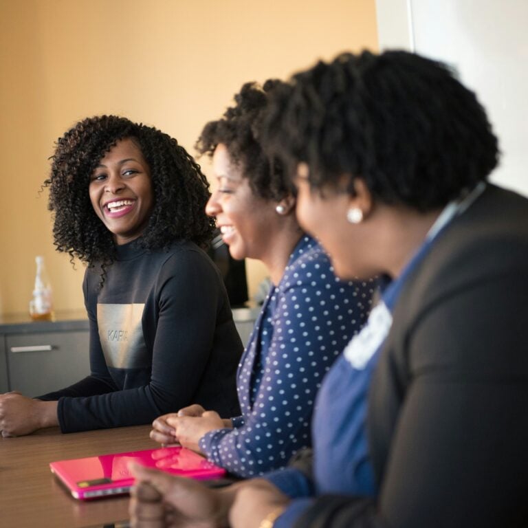 Three women sit together at a table, smiling and talking. One woman has a bright pink laptop in front of her. The atmosphere appears friendly and collaborative.