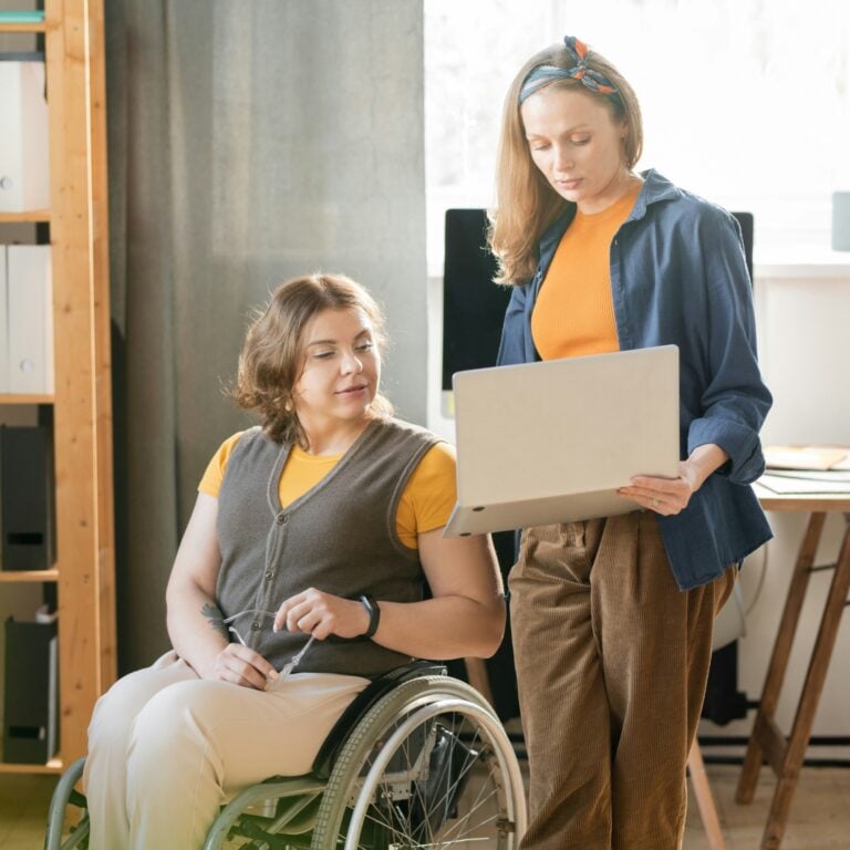 Two women in an office setting, one seated in a wheelchair holding spectacles, and the other standing beside her holding an open laptop. They are having a conversation and appear to be collaborating on work.