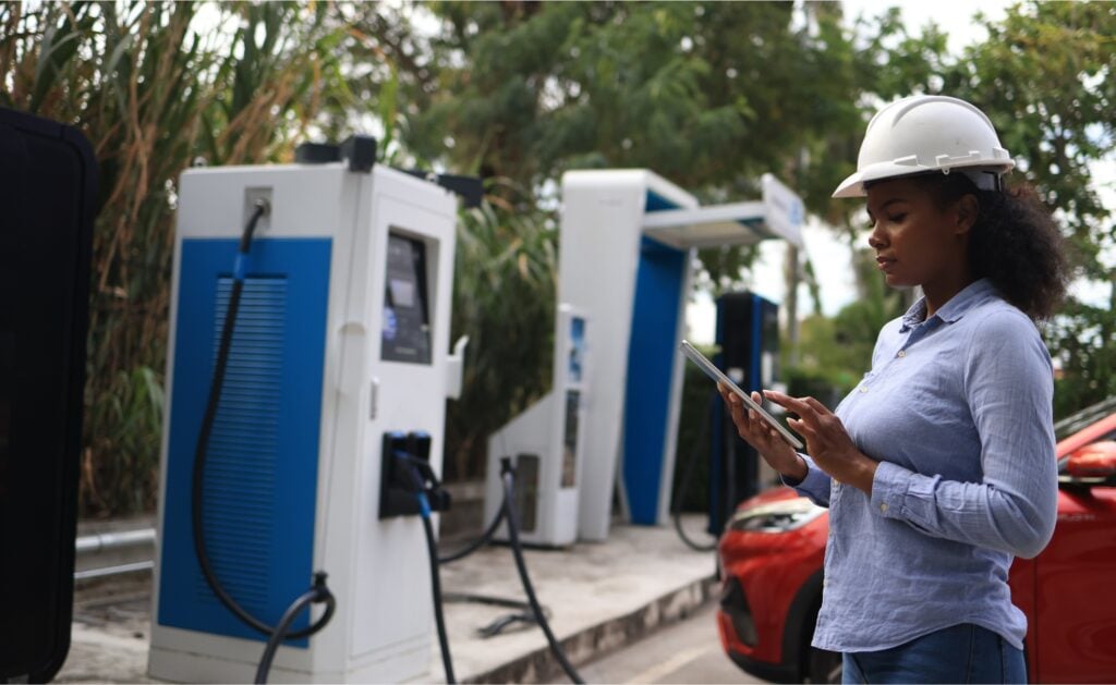 A woman wearing a white hard hat and blue shirt uses a tablet at an electric vehicle charging station outdoors, with a red car charging nearby and greenery in the background.