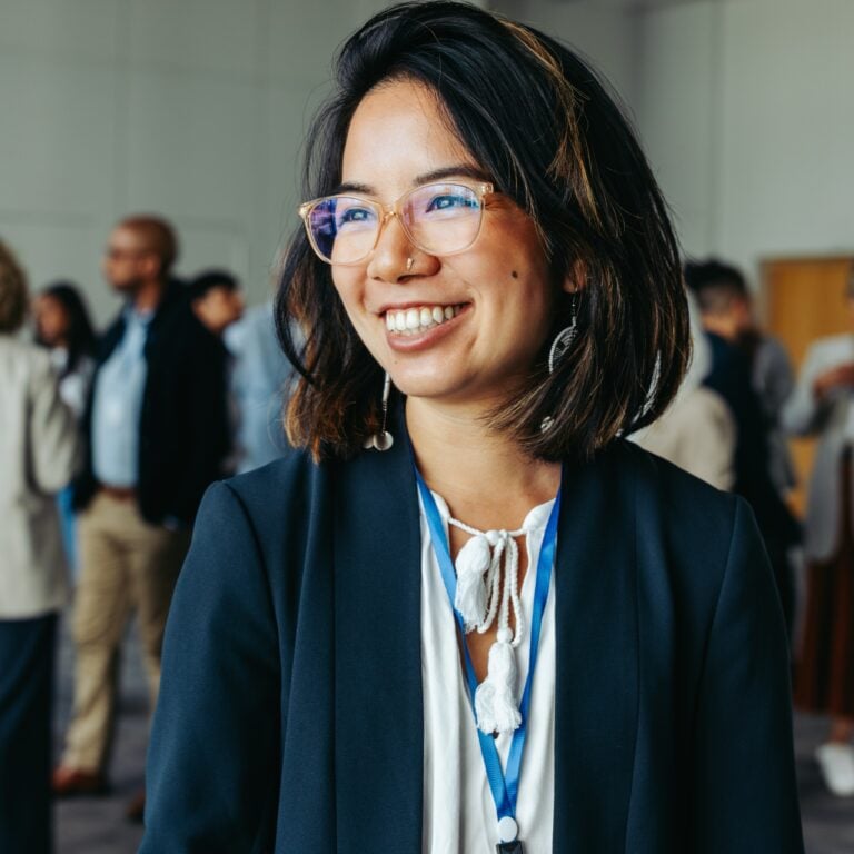 A smiling woman wearing glasses and a business outfit stands in a conference room with a lanyard around her neck. Other people are seen networking and talking in the blurred background.