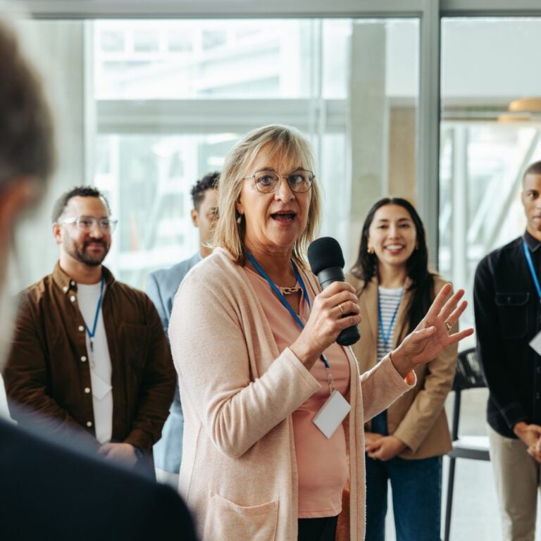 A woman holding a microphone speaks to a group of people, who are standing in a bright room and listening attentively. Some of the listeners are smiling and wearing name badges.